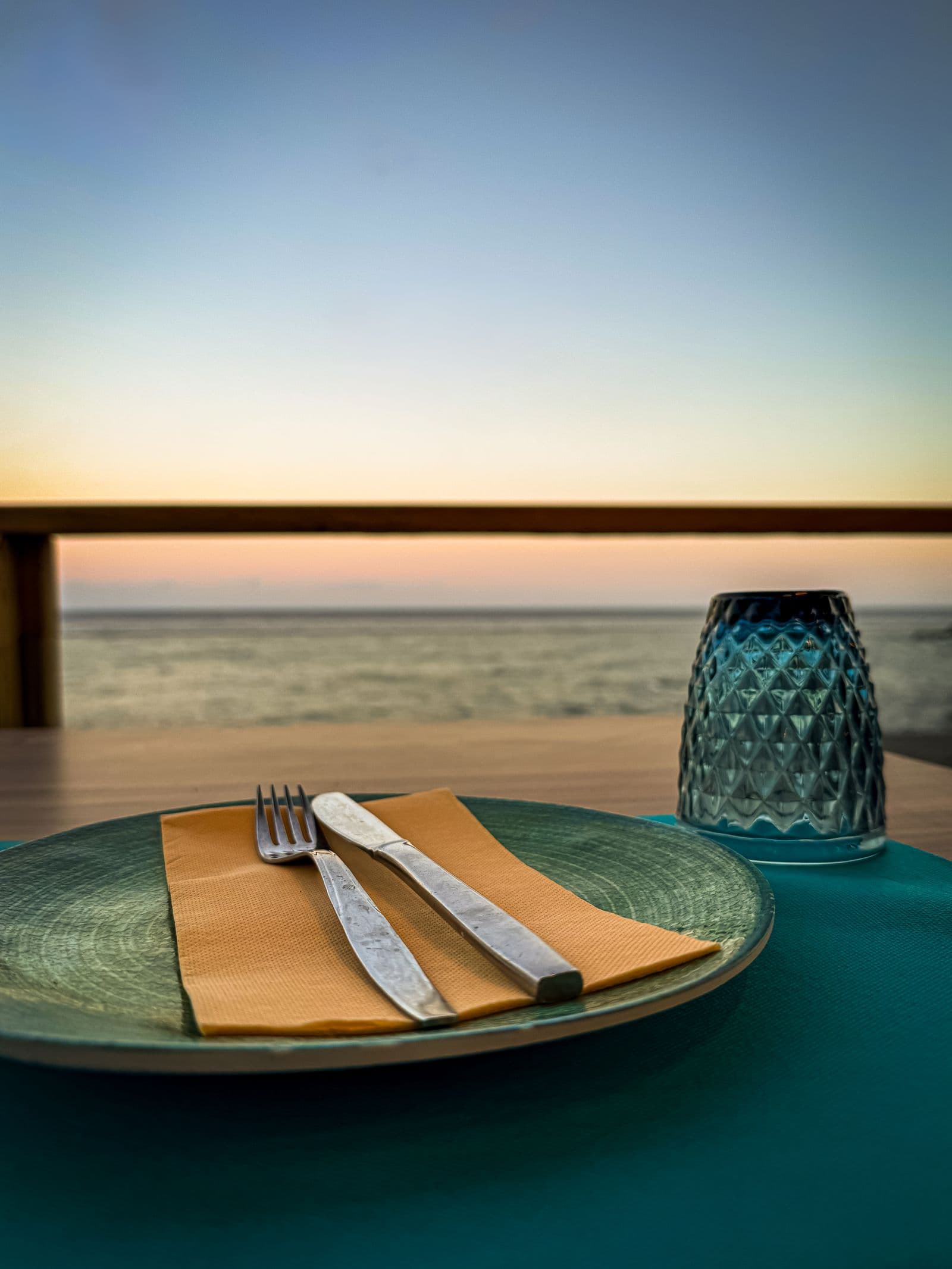 A dinner place setting on a terrace, the sea and a low sun behind it